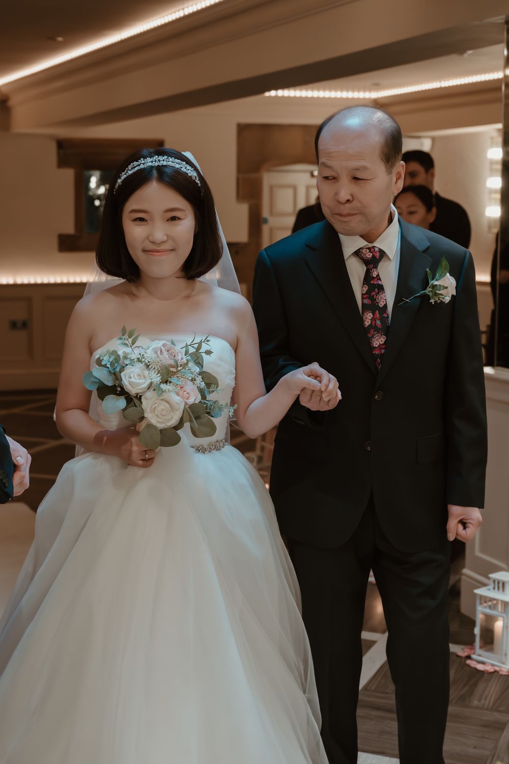 Bride in white gown holding bouquet walks down aisle with older man in dark suit at indoor wedding ceremony.