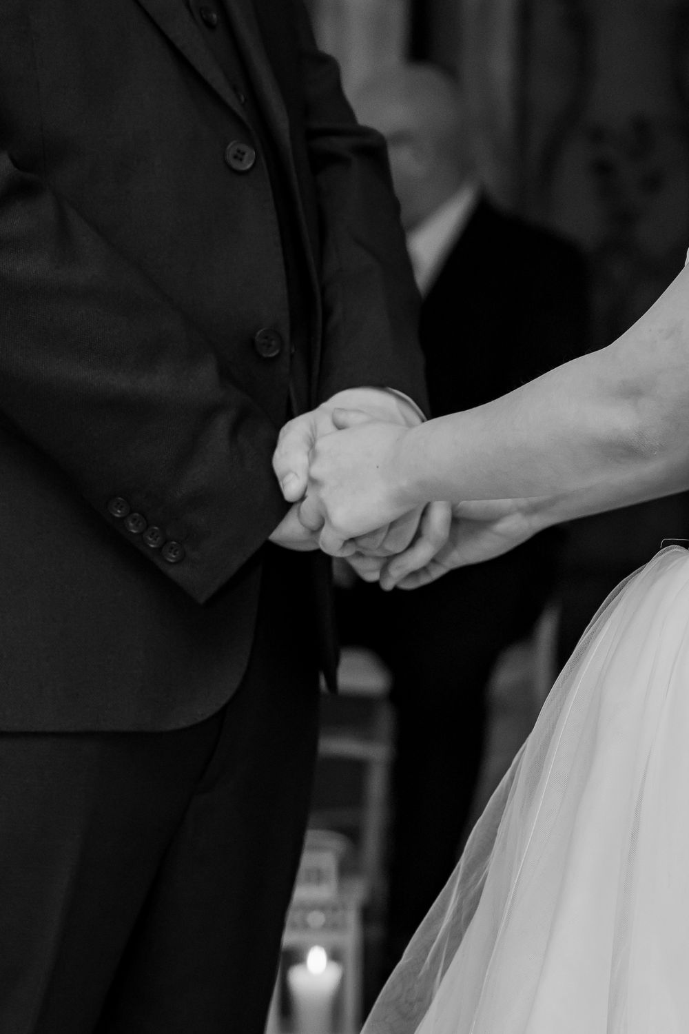 Black and white photo of bride and groom holding hands at wedding ceremony with candle in background.