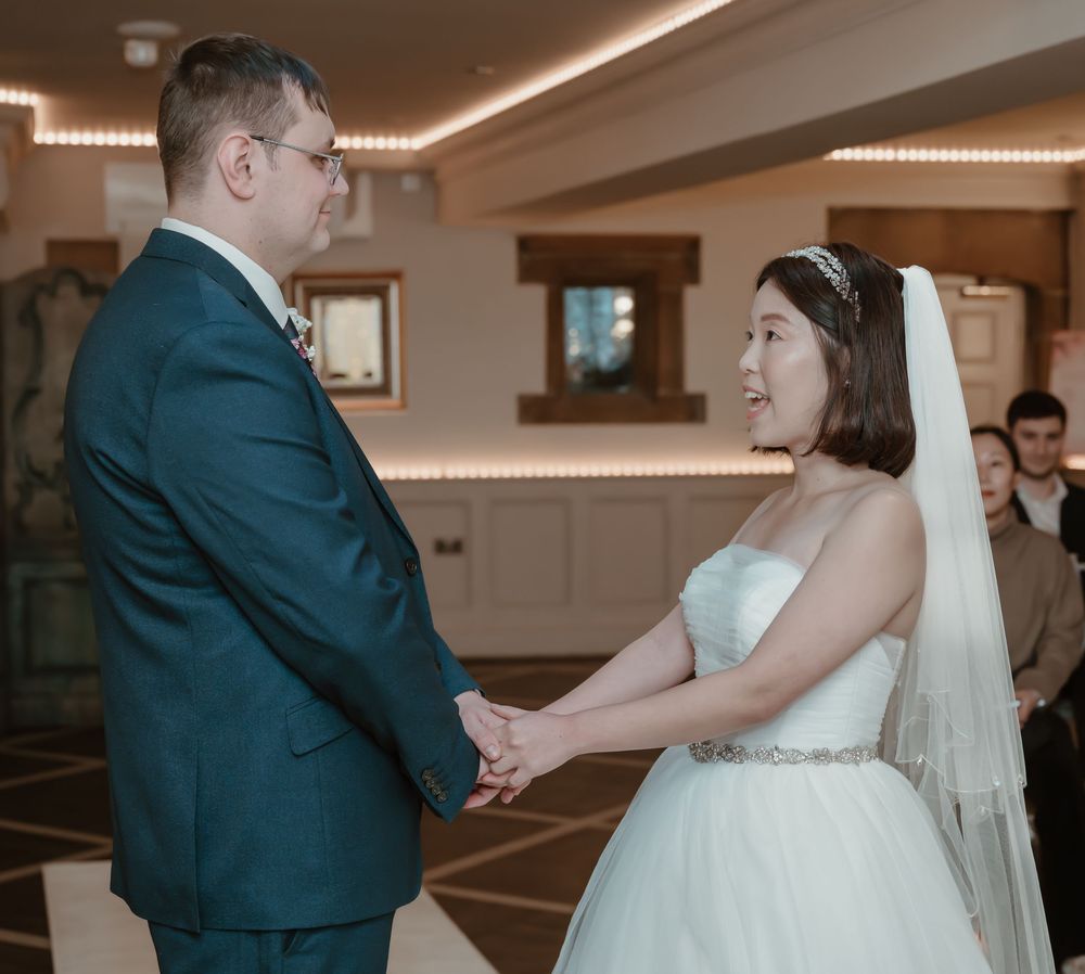 A couple exchanges vows at their wedding ceremony, the groom in a teal suit holding hands with the bride in a white gown.