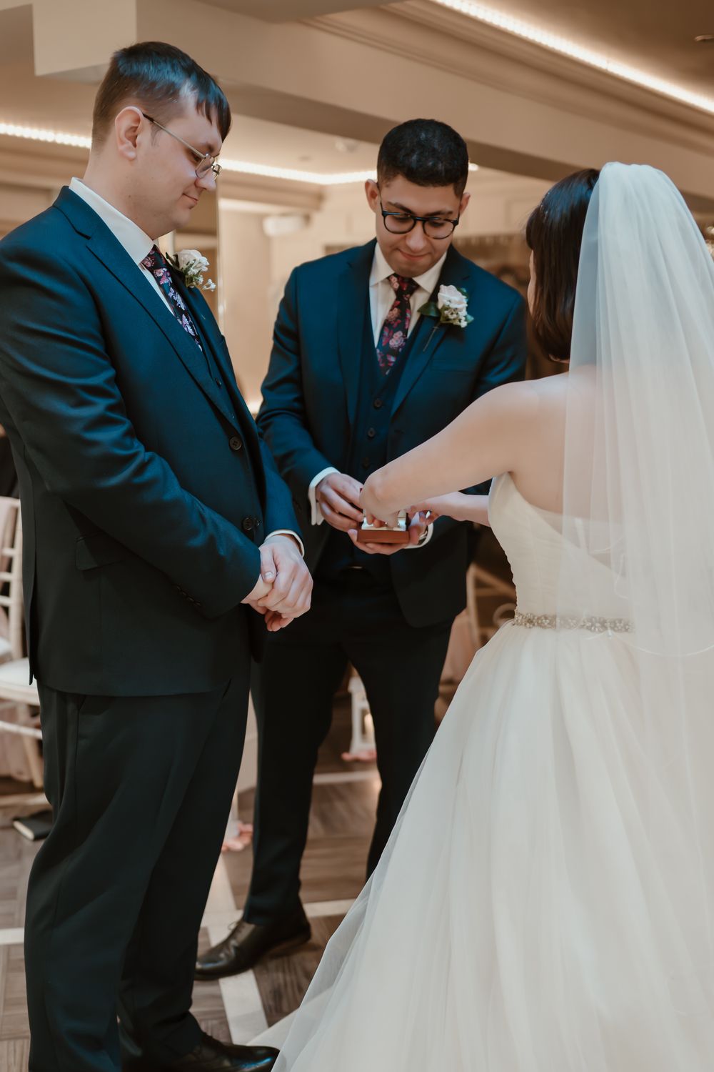 Bride placing wedding ring on groom's finger during indoor ceremony, officiant watching, both men in navy suits.
