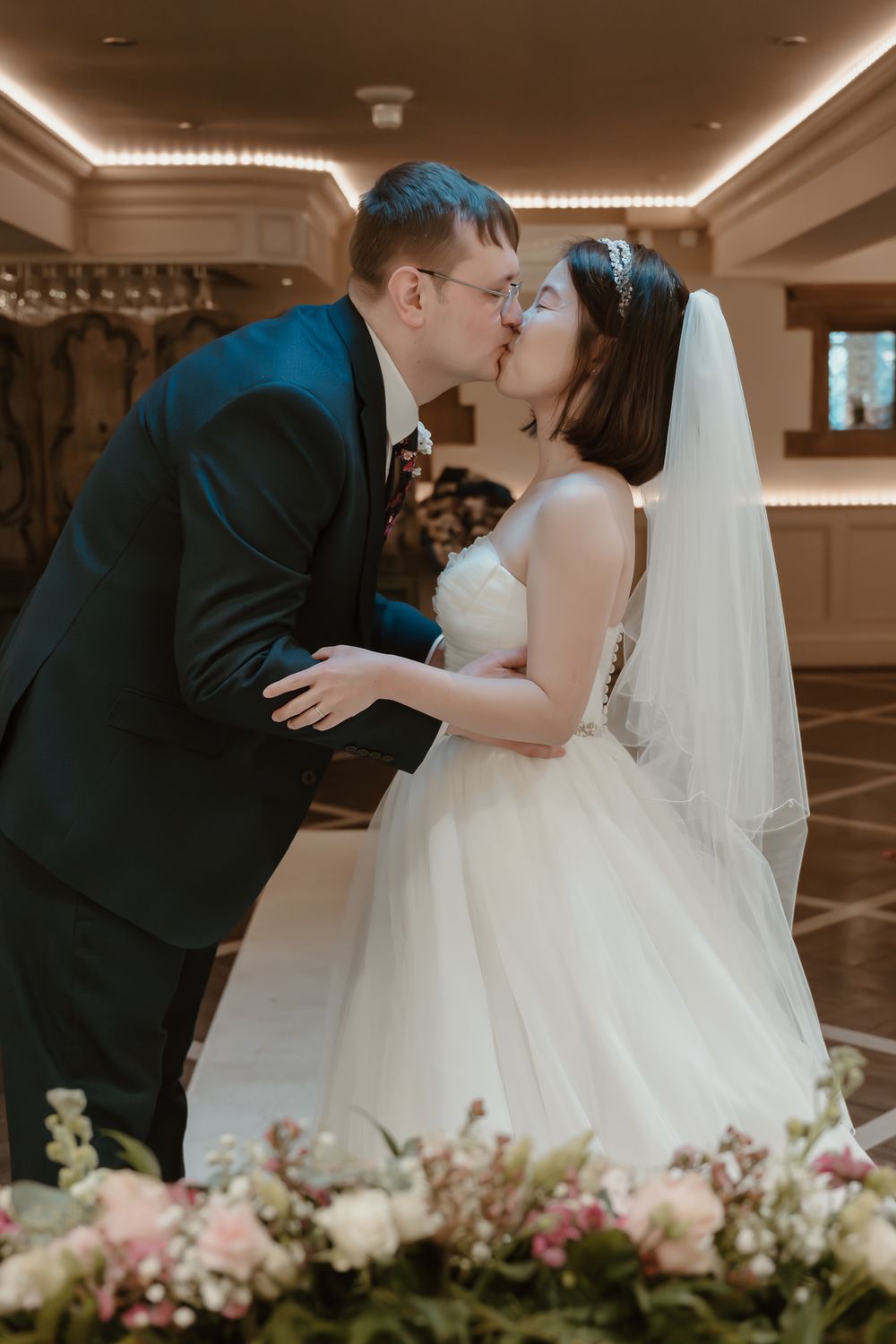 Bride and groom share their first kiss at wedding ceremony, bride in white tulle gown with veil, groom in dark suit.