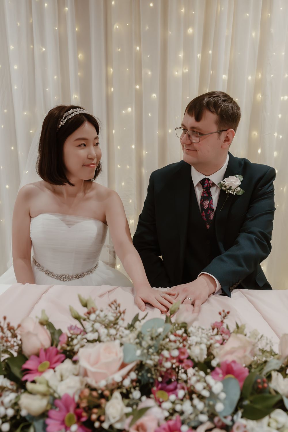 A bride and groom sit together at a floral-decorated table, smiling at each other before a twinkling fairy light backdrop.