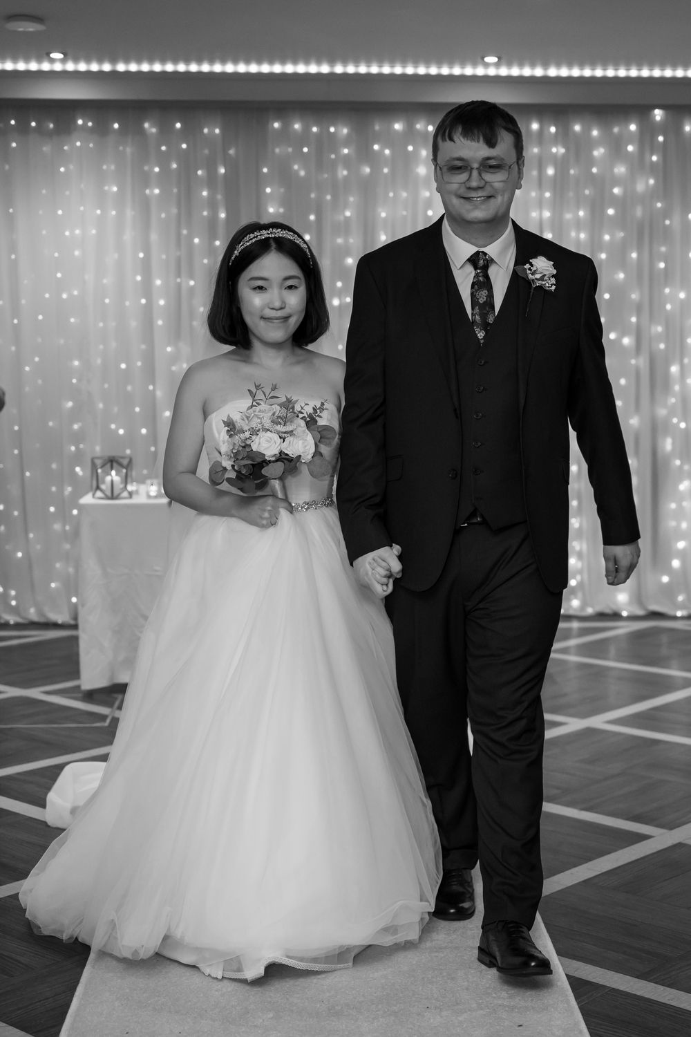 Black and white photo of a bride holding flowers and groom in suit walking together at wedding reception.