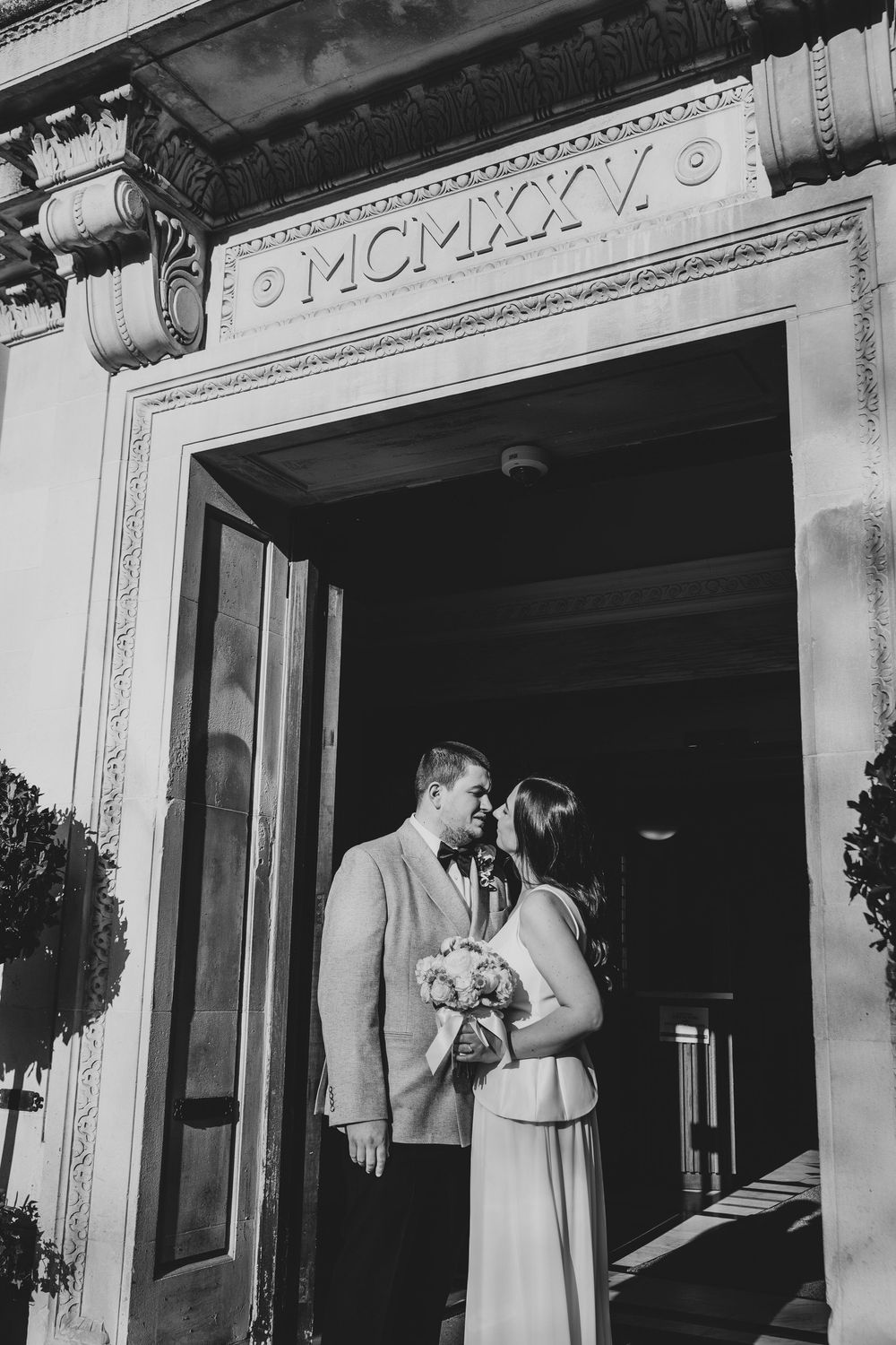 Black and white photo of a couple kissing in a doorway, bride holding bouquet, building inscribed MCMXXXV.