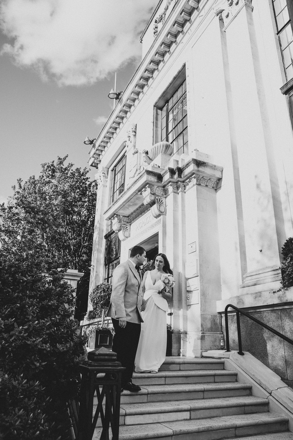 Black and white photo of a bride and groom standing on steps outside an elegant classical building on their wedding day.