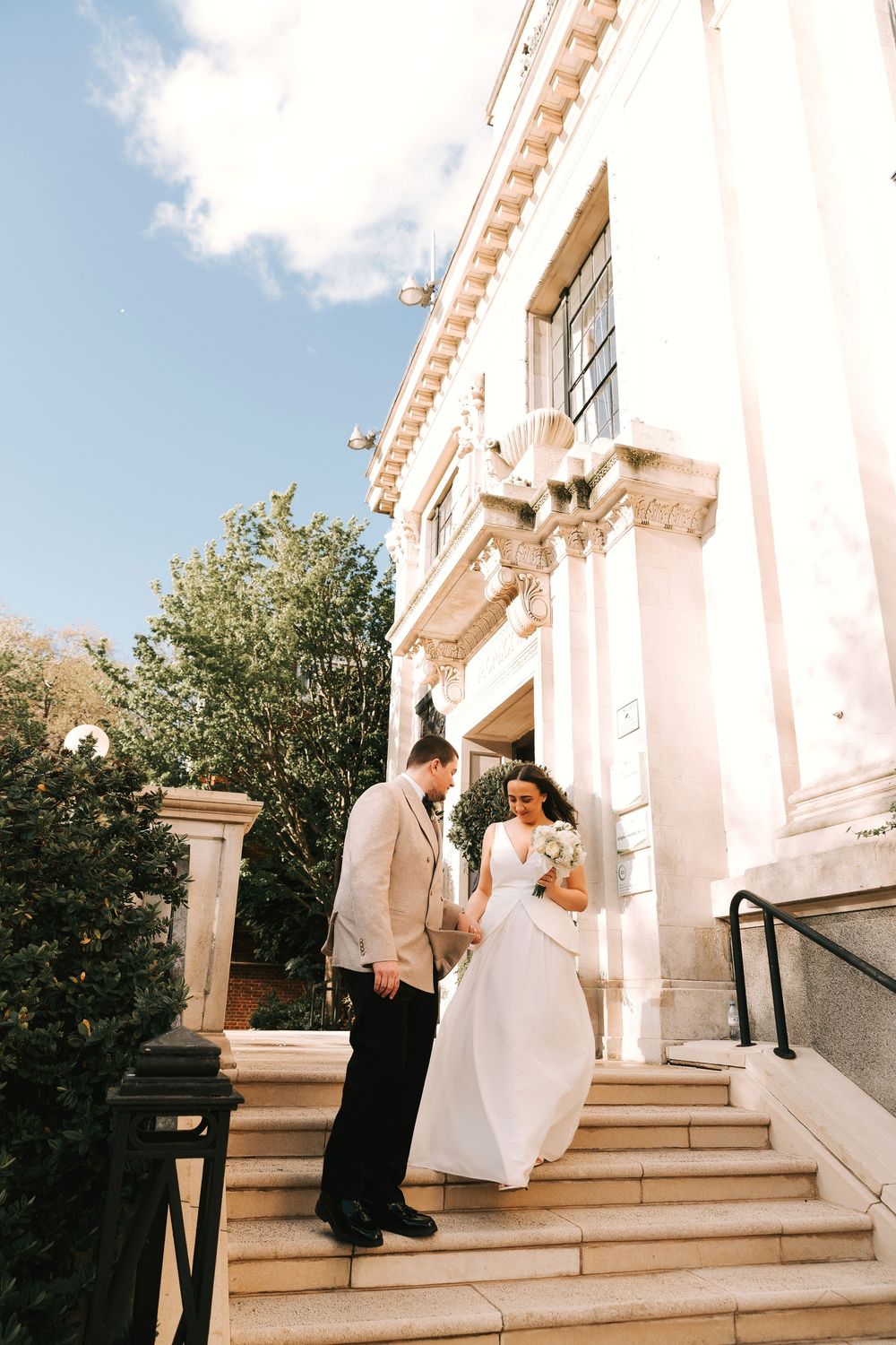 Bride and groom descend stone steps of elegant classical building on sunny wedding day.