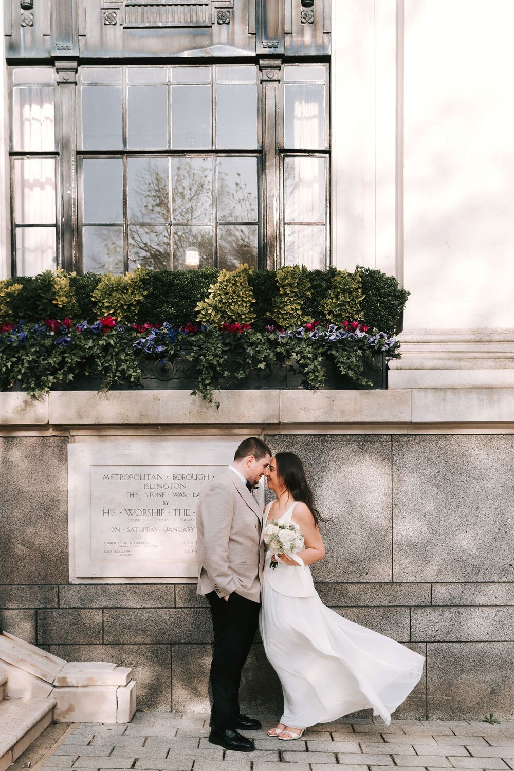 Bride and groom share a romantic moment outside a classic stone building with lush flower boxes above.