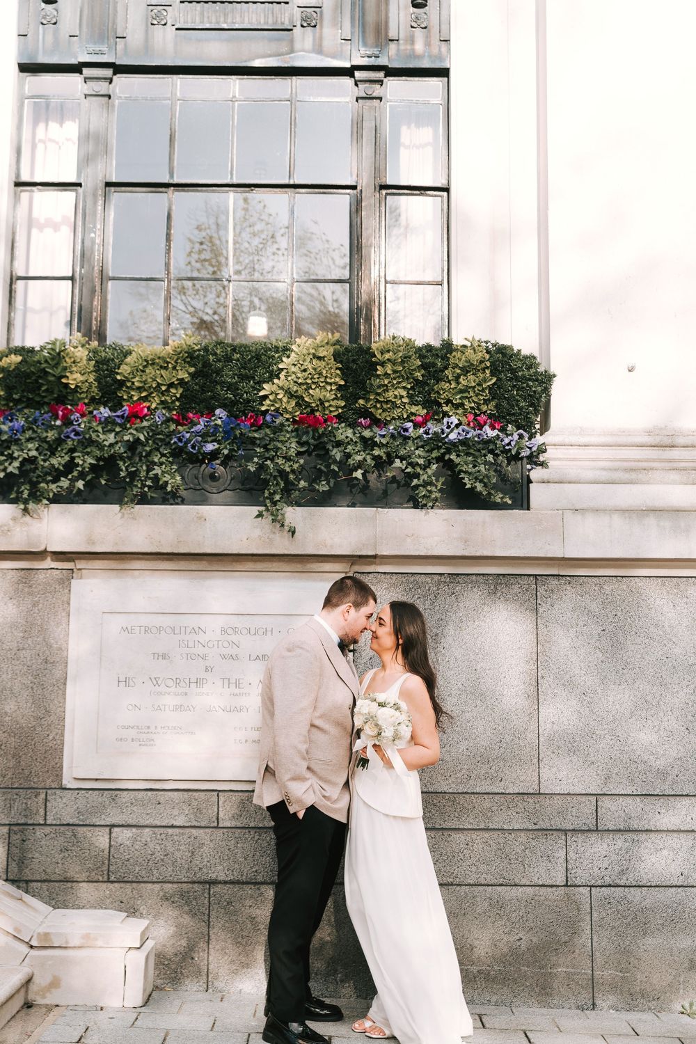 Bride and groom share a romantic moment outside a stone building with a floral window box backdrop.