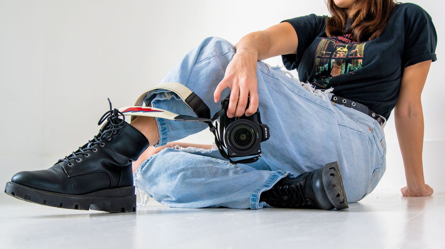Photographer sitting on floor in ripped jeans and black boots, holding a DSLR camera against white background.