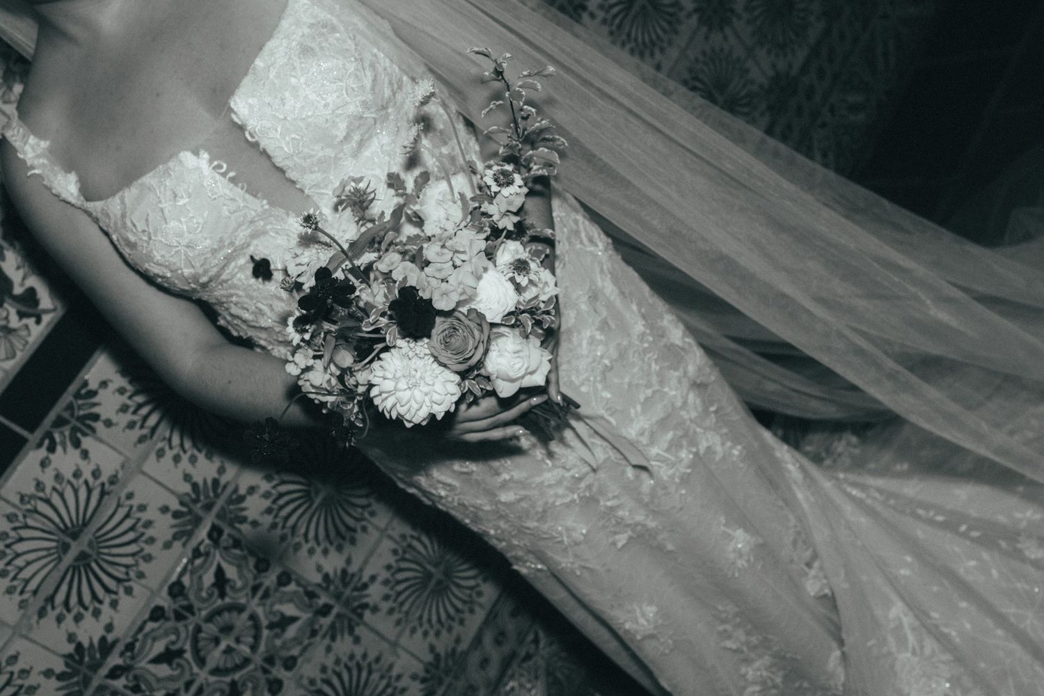 Black and white photo of a bride in lace wedding dress holding a floral bouquet with veil draped elegantly.
