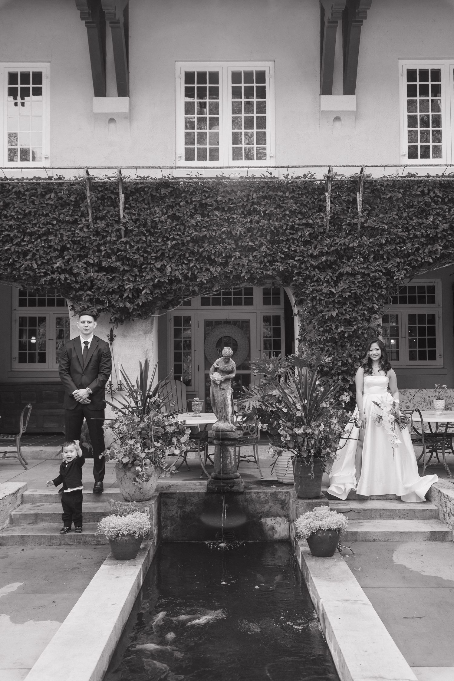 Black and white wedding photo of bride and groom by ivy-covered mansion with fountain courtyard.