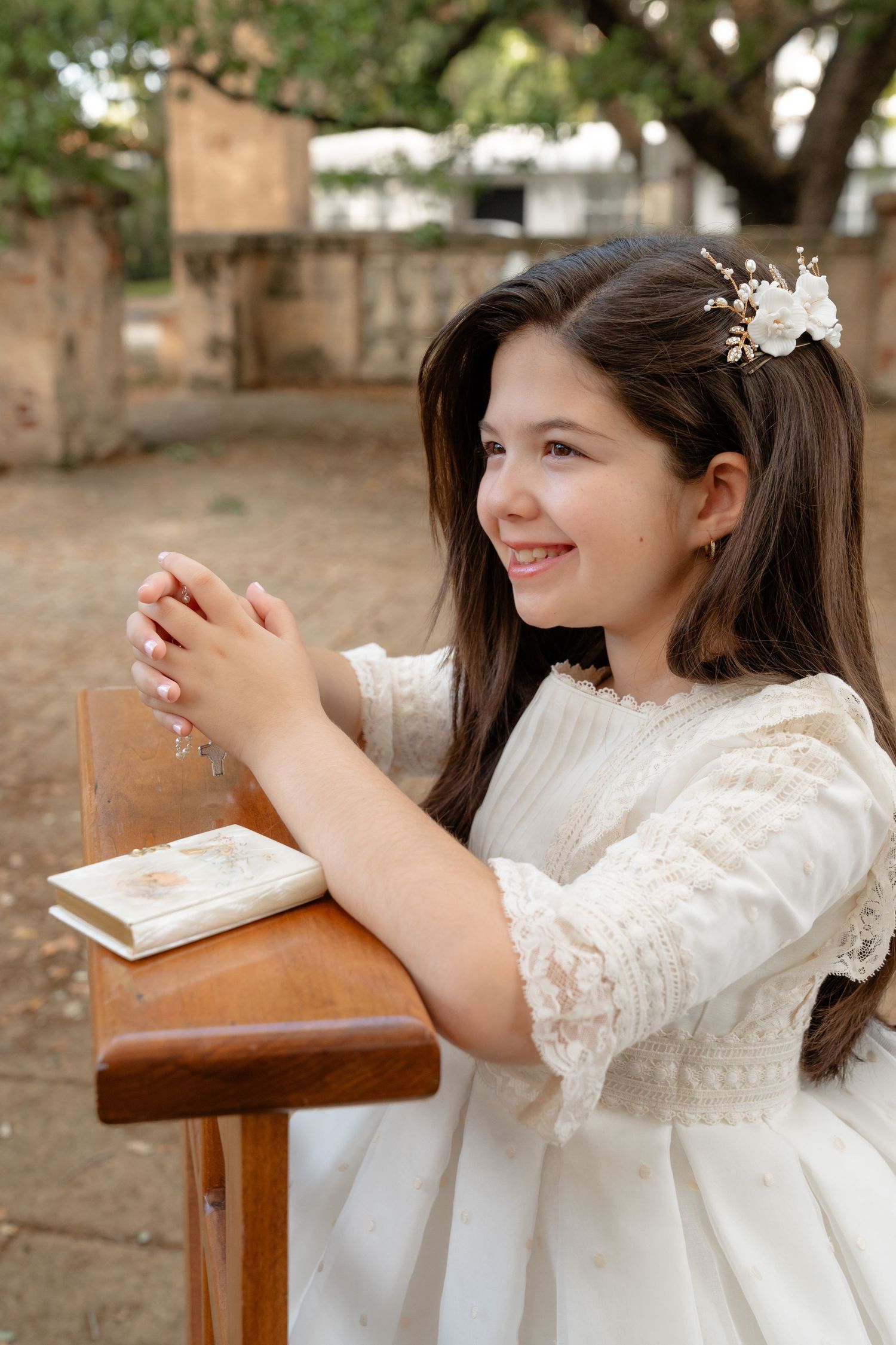 Young girl in white lace dress holding rosary beads at wooden prie-dieu, smiling during First Communion ceremony.