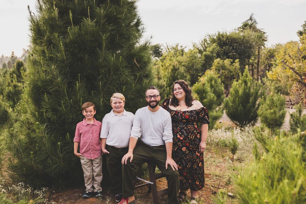 Family of four smiling together among tall green pine trees outdoors during a mini session with heidi grace photography