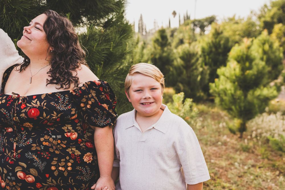 A woman and young boy smile and laugh together outdoors among green pine trees during a family photo session near redlands ca