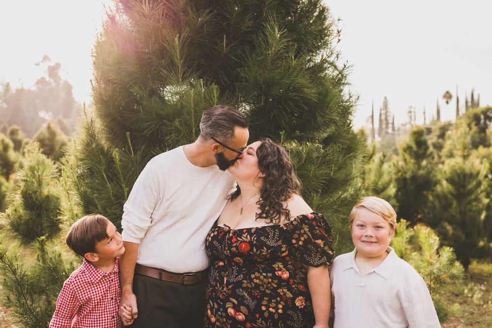 Family kissing at Christmas tree farm mini session with two young boys smiling outdoors in warm sunlight in the inland empire