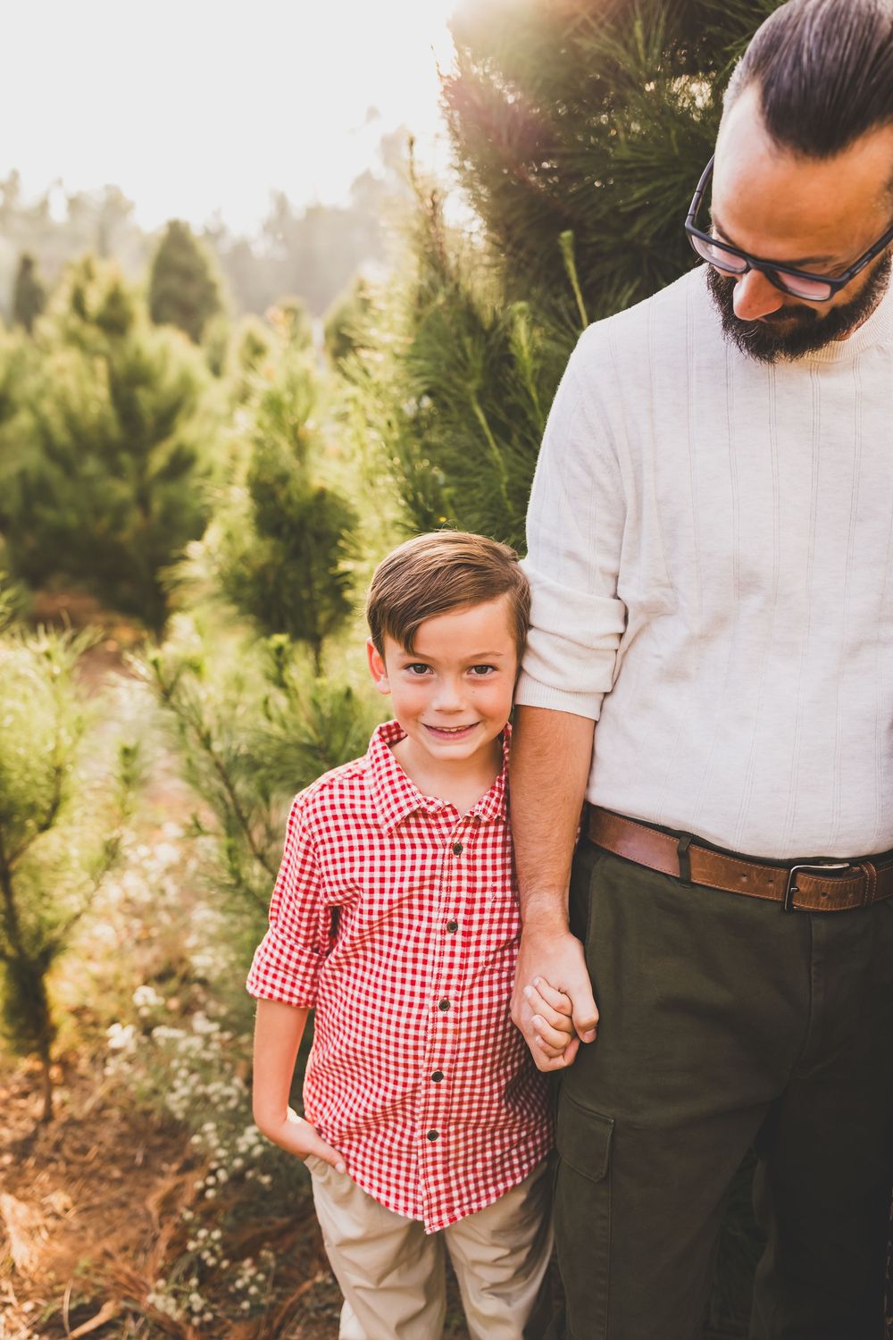 A smiling boy holds hands with a bearded man among green pine trees at a Christmas tree farm with heidi grace photography