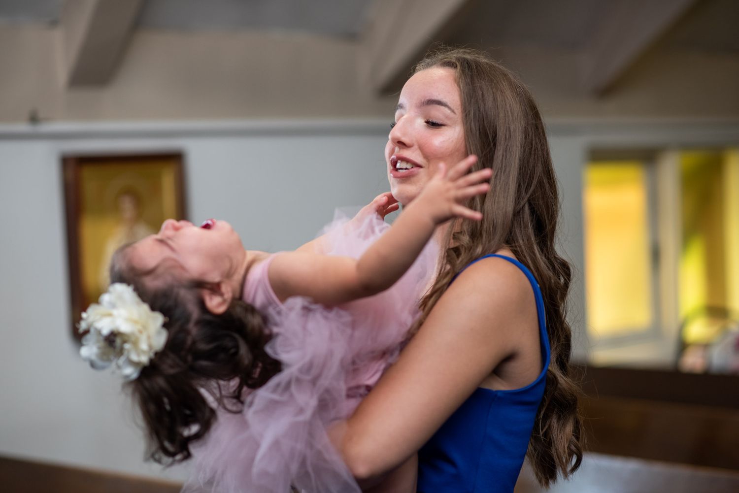Young teenage girl in blue dress joyfully holding a laughing toddler in pink tutu in church.
