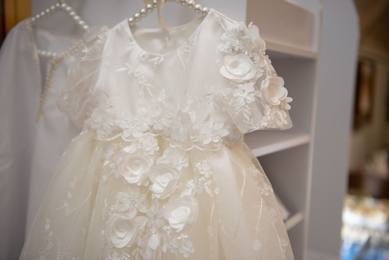 Elegant ivory baptism dress with 3D floral appliques and tulle skirt hanging on a white wardrobe.