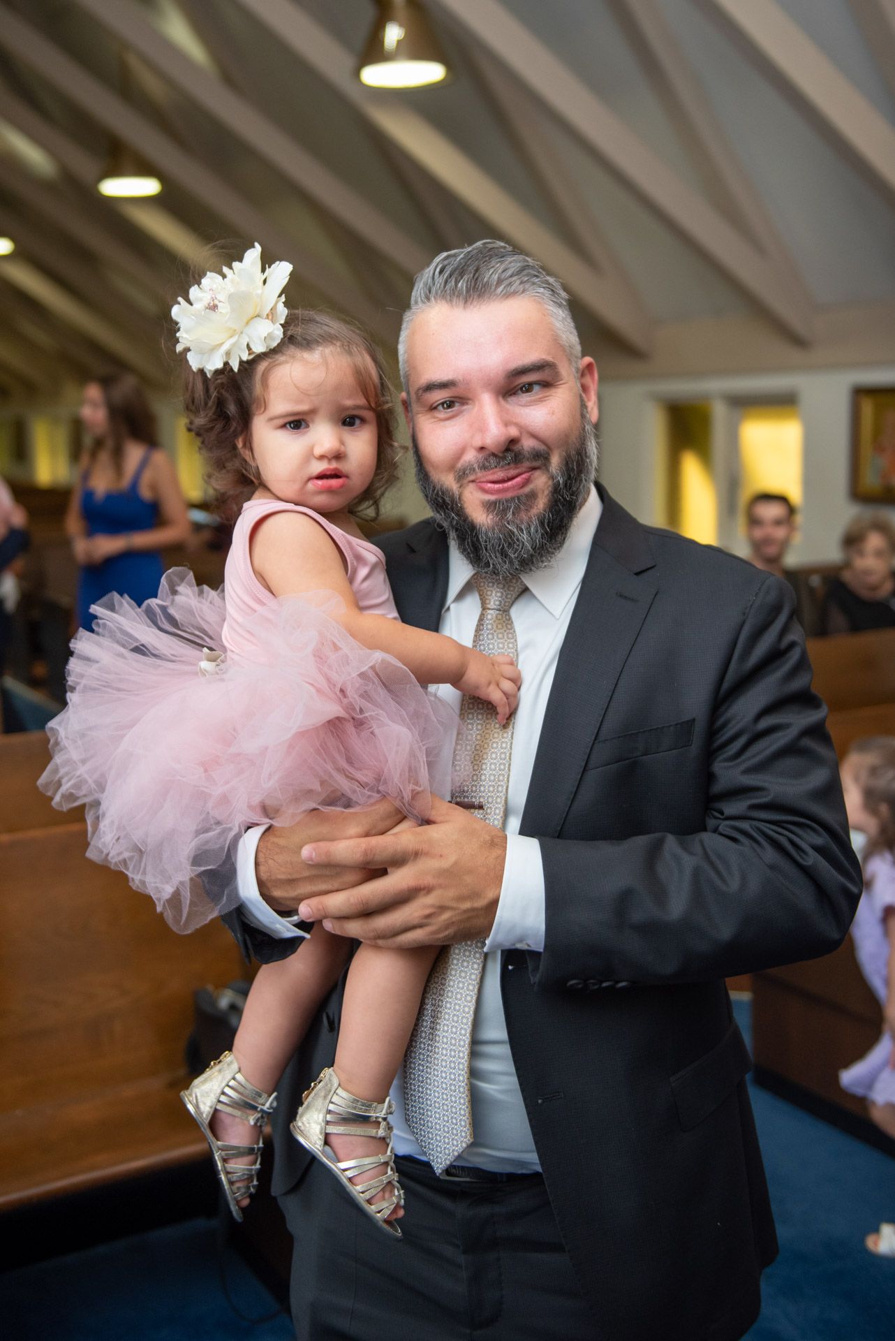 Smiling bearded man in suit holding a toddler girl in pink tutu dress inside a church.
