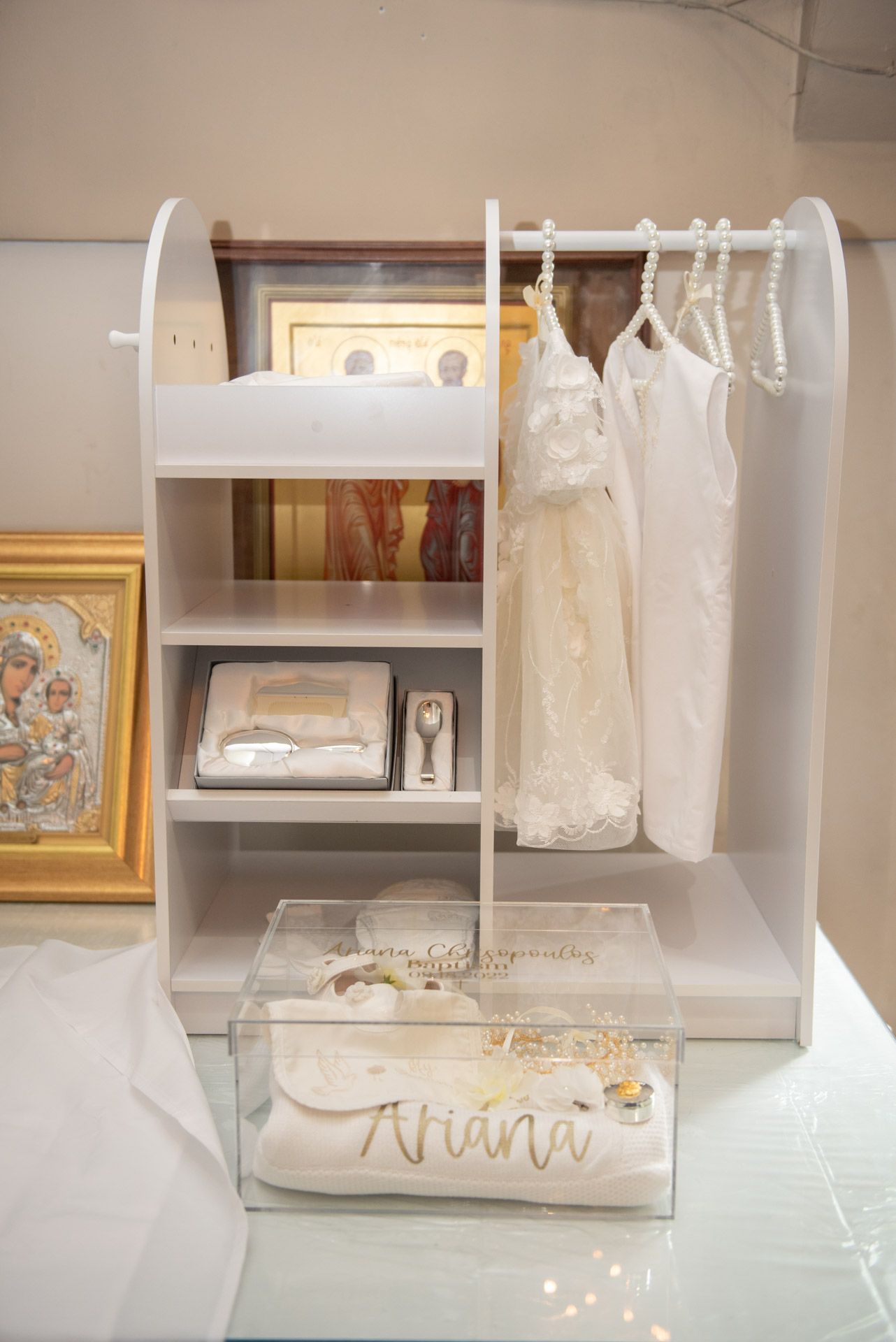 White baptism display unit with shelves holding keepsakes, white gown, and personalized Ariana box.