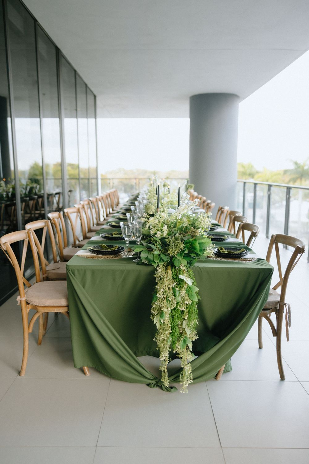 Elegant wedding reception table with green tablecloth, floral garland centerpiece, wooden cross-back chairs on a modern venue balcony.