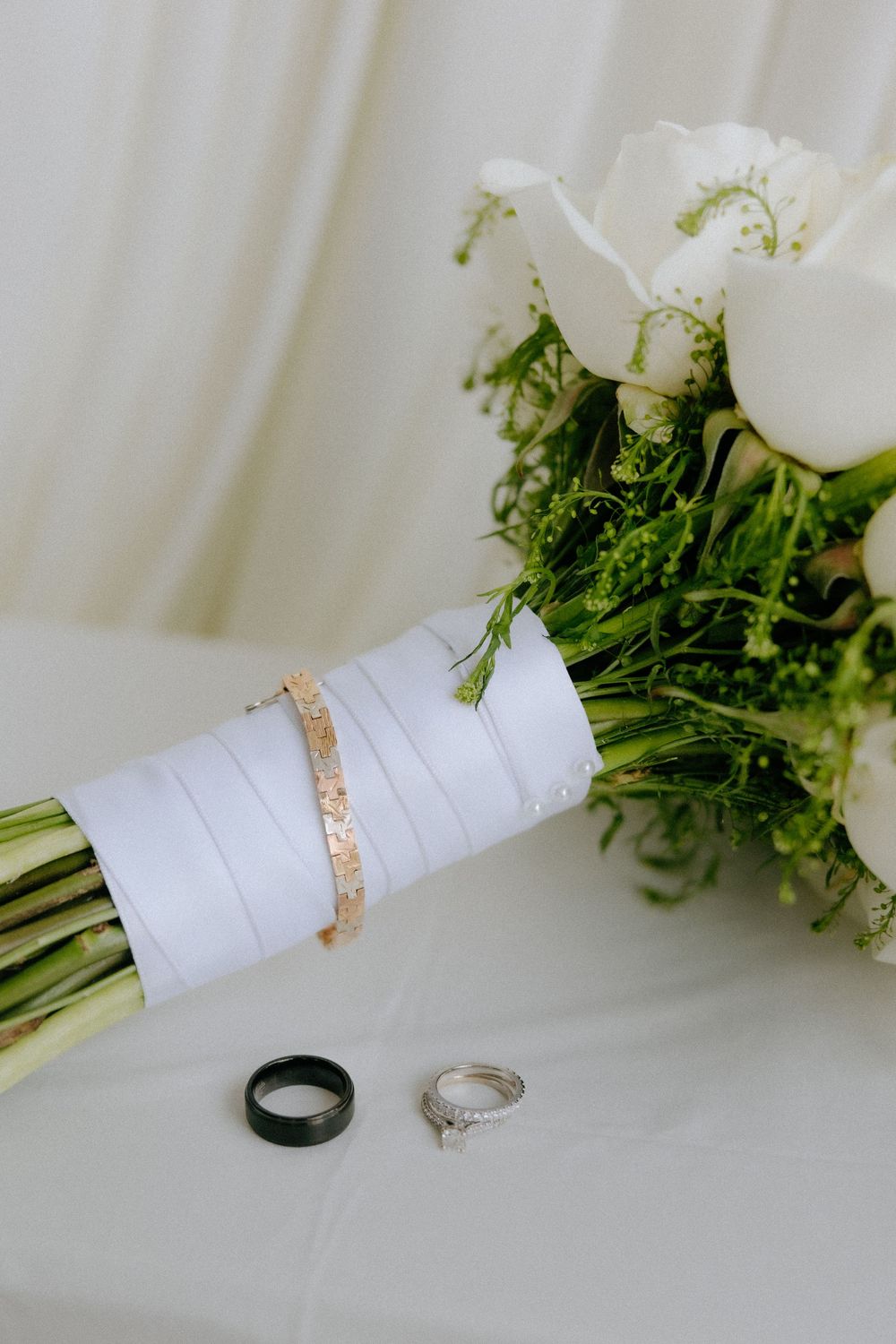 Wedding bouquet of white roses with rings and bracelet displayed on white fabric.