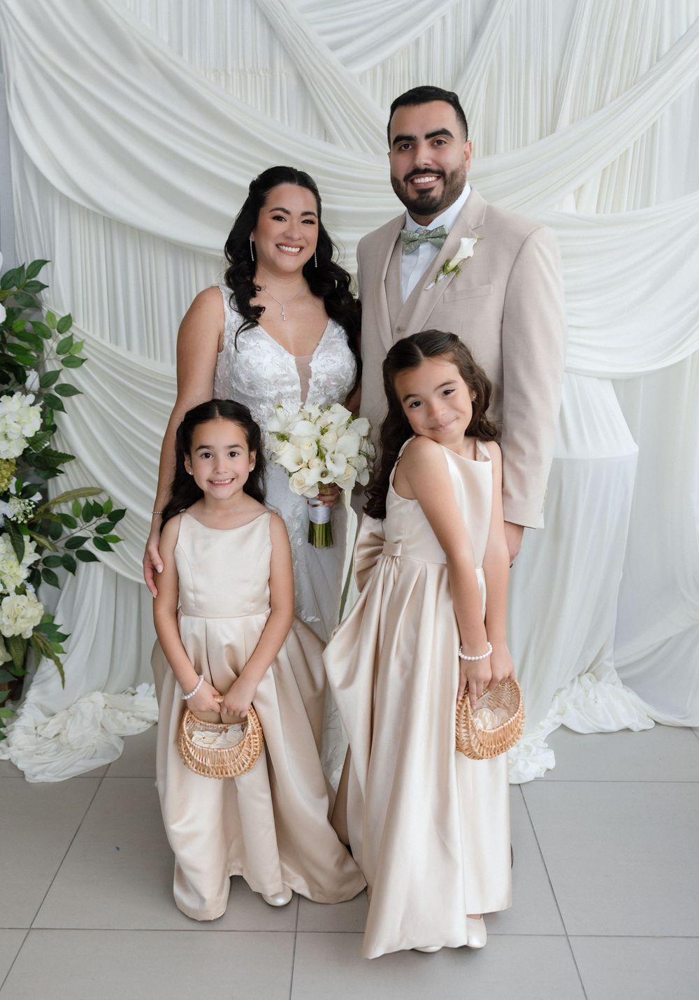 Bride and groom pose with two flower girls in champagne dresses before a white draped backdrop at an elegant wedding.