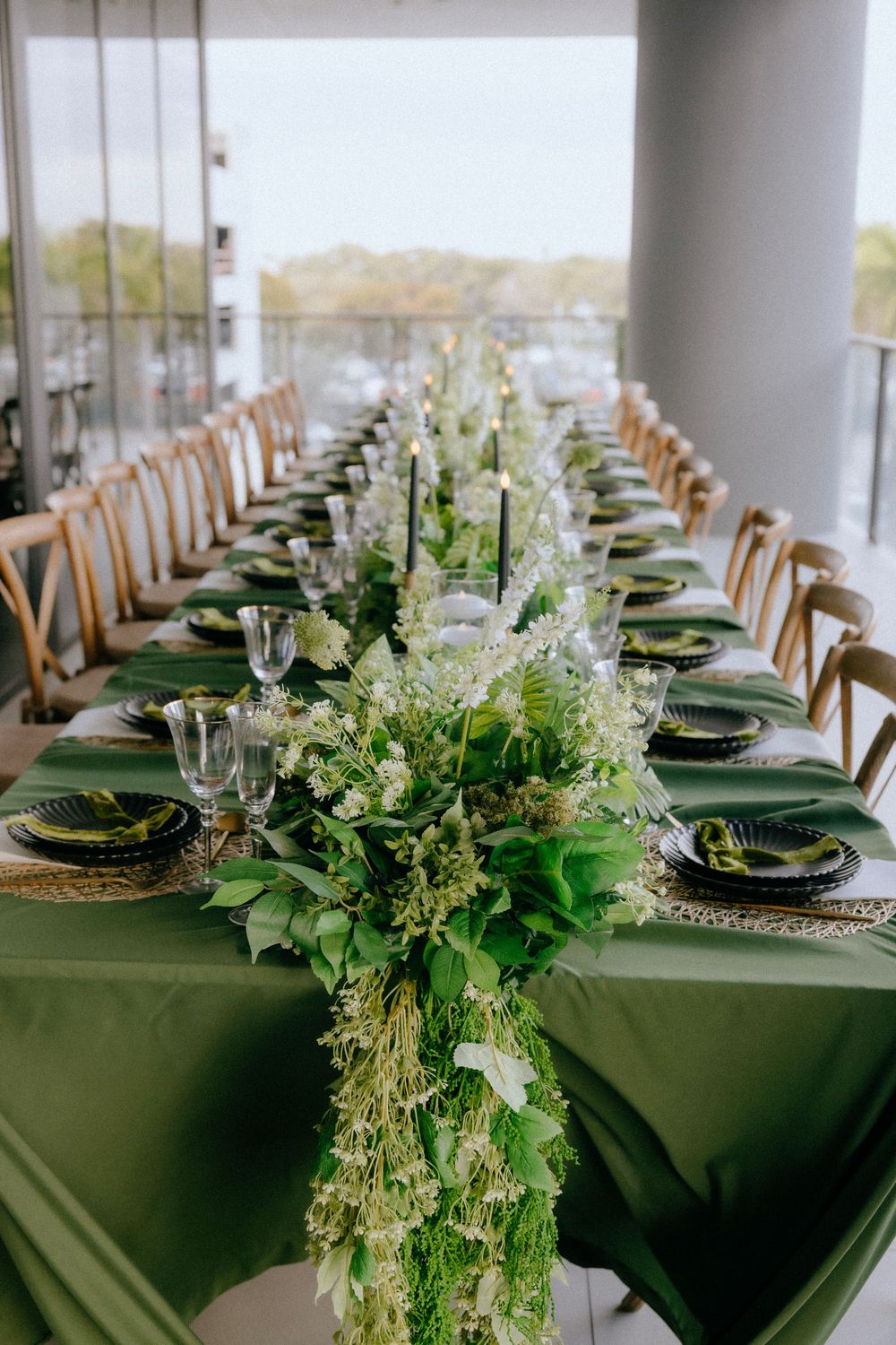 Elegant long dining table with green linen, floral garland centerpiece, black plates, and gold chargers for a formal event.