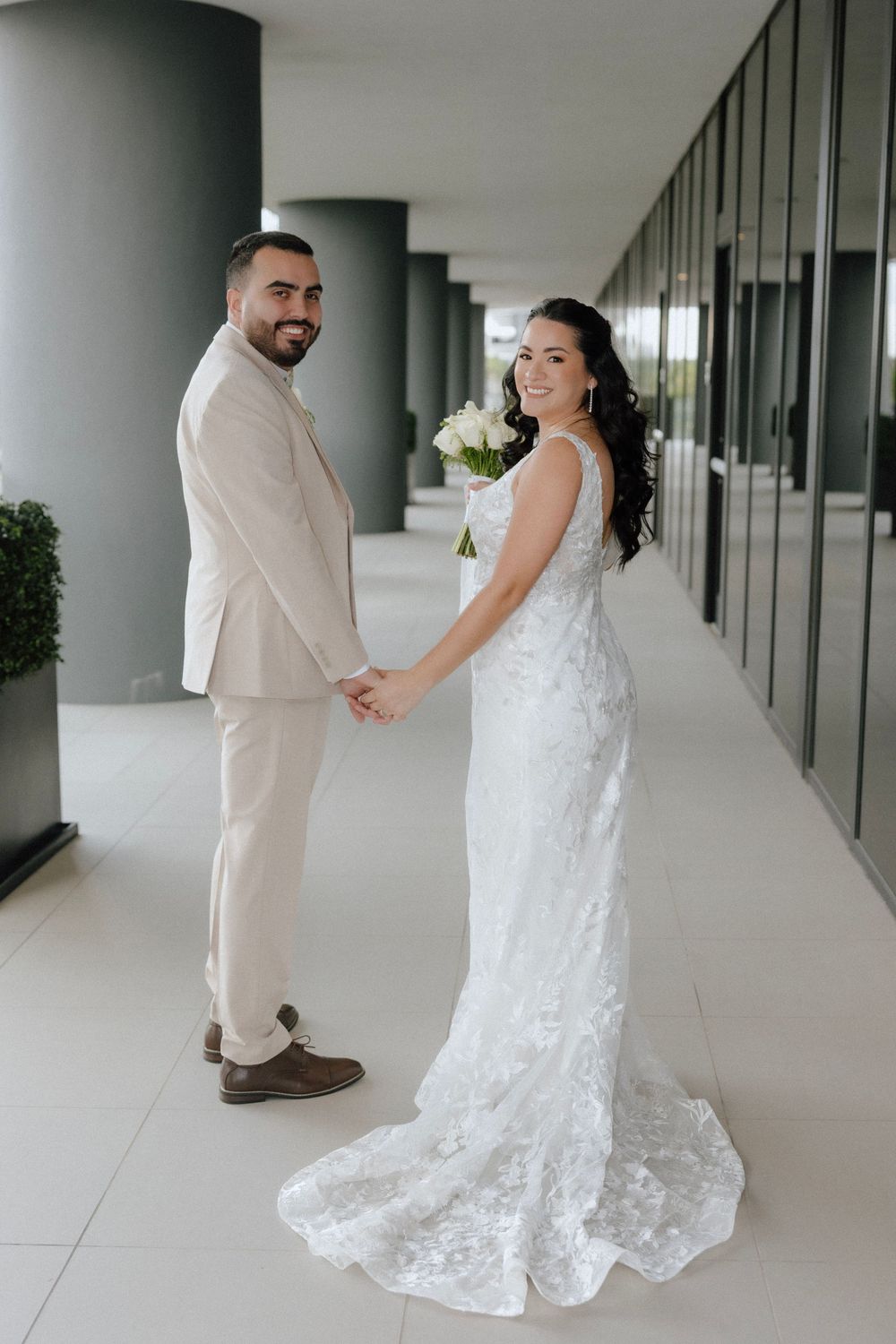 Bride in lace gown holding bouquet and groom in beige suit hold hands, smiling at camera in modern venue hallway.