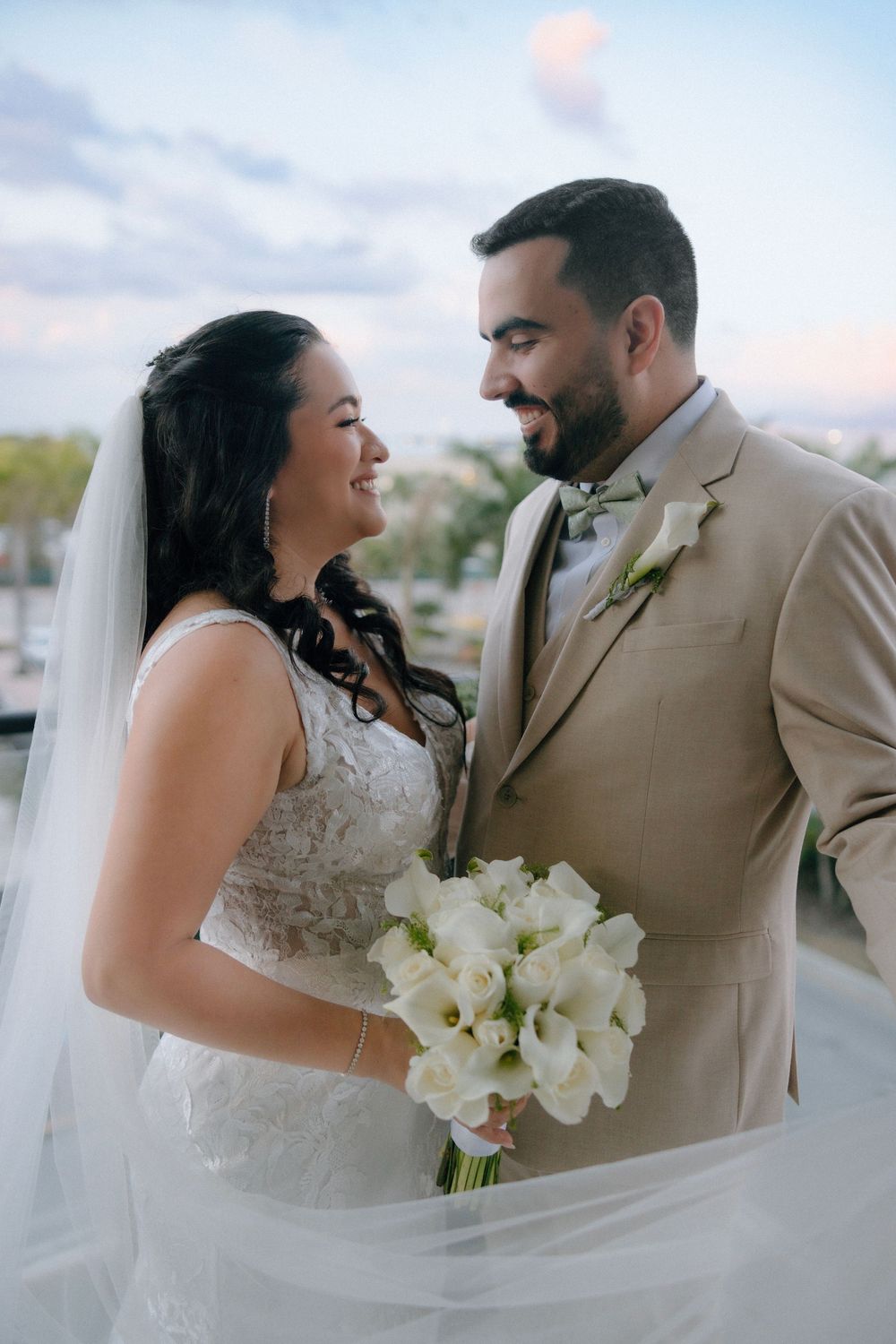 Bride and groom share a romantic moment outdoors, bride holding white calla lily bouquet, groom in tan suit.