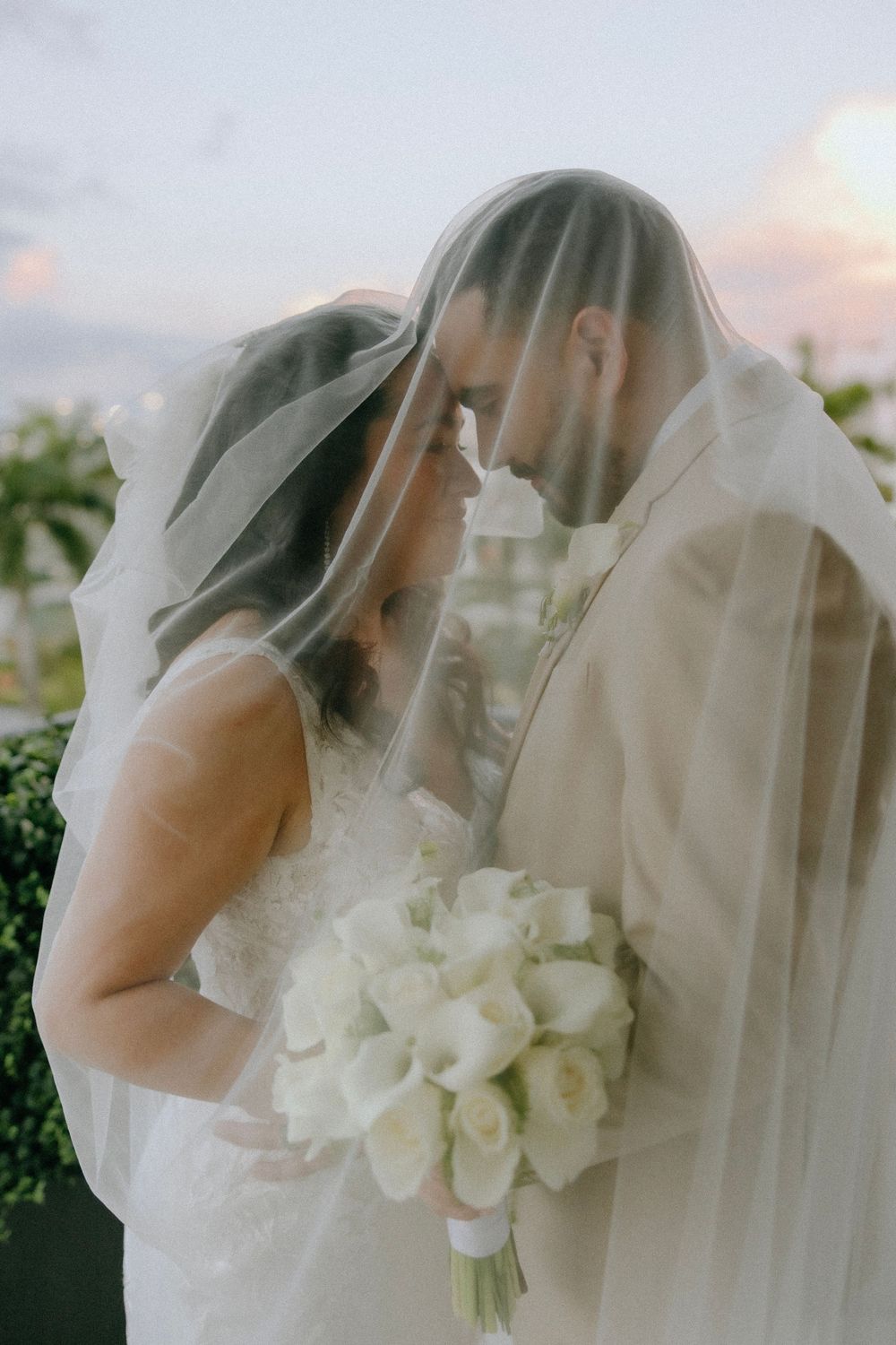 Bride and groom share a romantic moment under a flowing white veil, bride holding white calla lily bouquet at sunset.