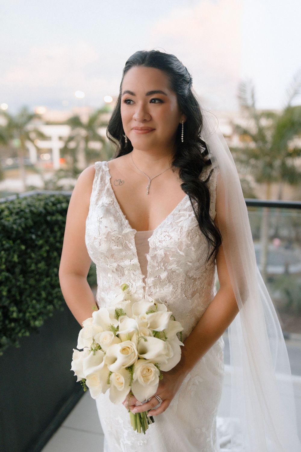 Bride in lace wedding gown and veil holds white calla lily bouquet on outdoor balcony with palm trees.