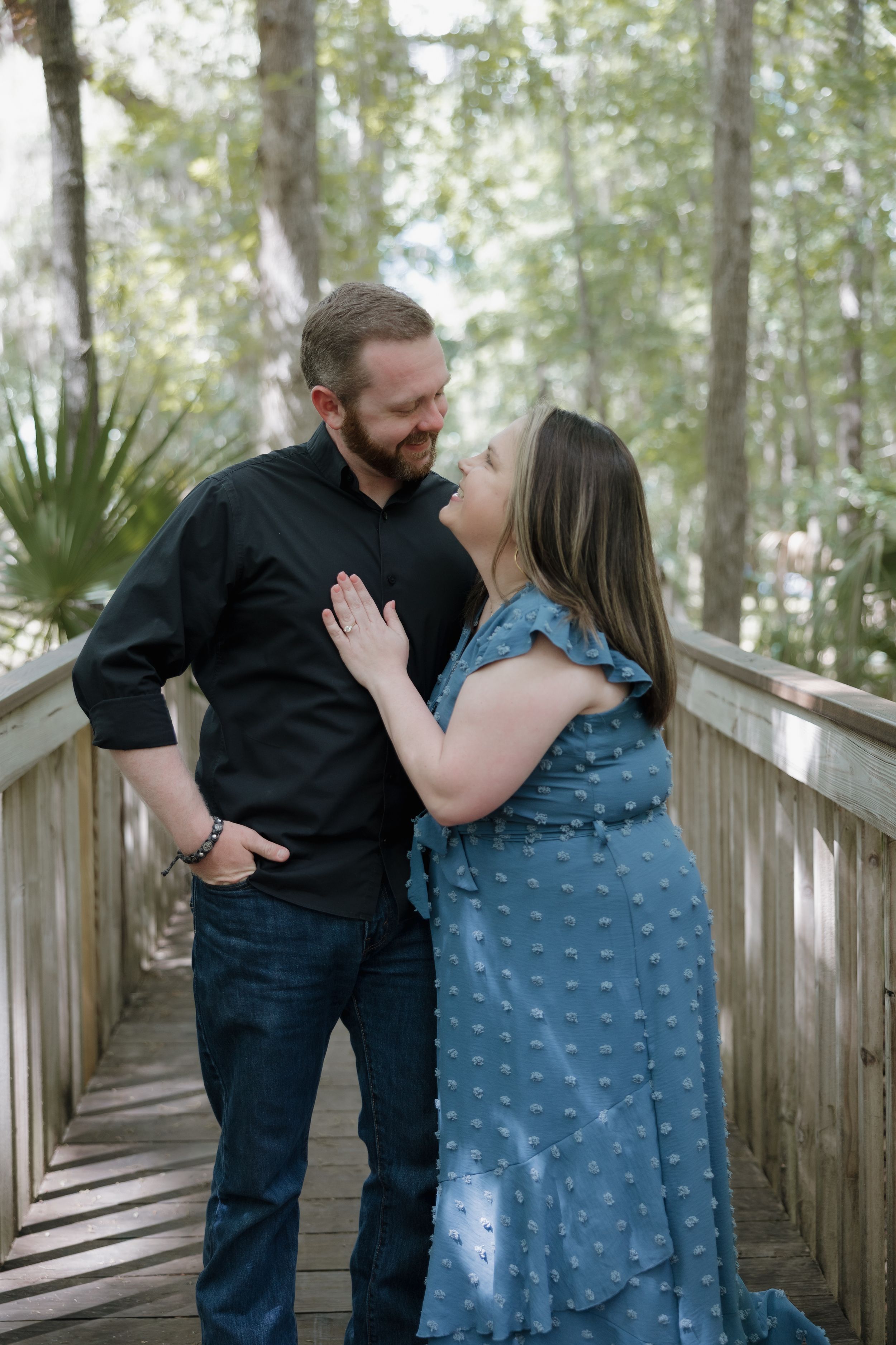 Couple sharing a romantic moment on a wooden bridge surrounded by lush green trees, woman in blue dress.