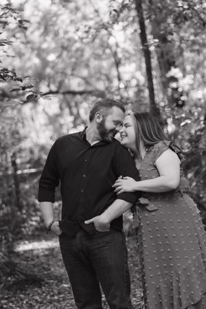 Black and white photo of a smiling couple embracing in a wooded setting, sharing a tender moment together.