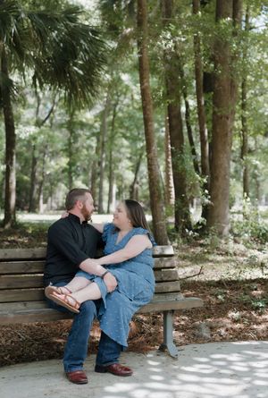 Couple sitting on a park bench holding hands, surrounded by tall trees, sharing a romantic moment outdoors.