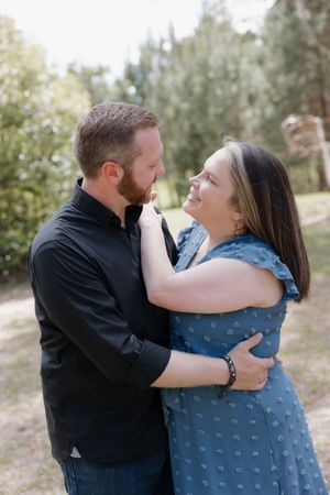 A couple embraces and gazes at each other lovingly outdoors, man in black shirt, woman in blue floral dress.