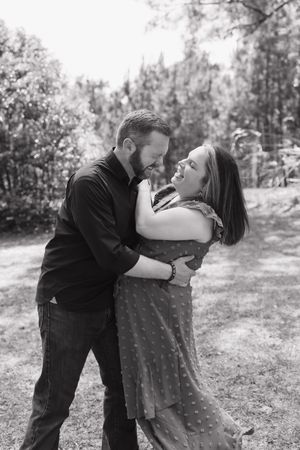 Black and white photo of a couple embracing and smiling at each other outdoors in a lush green park setting.