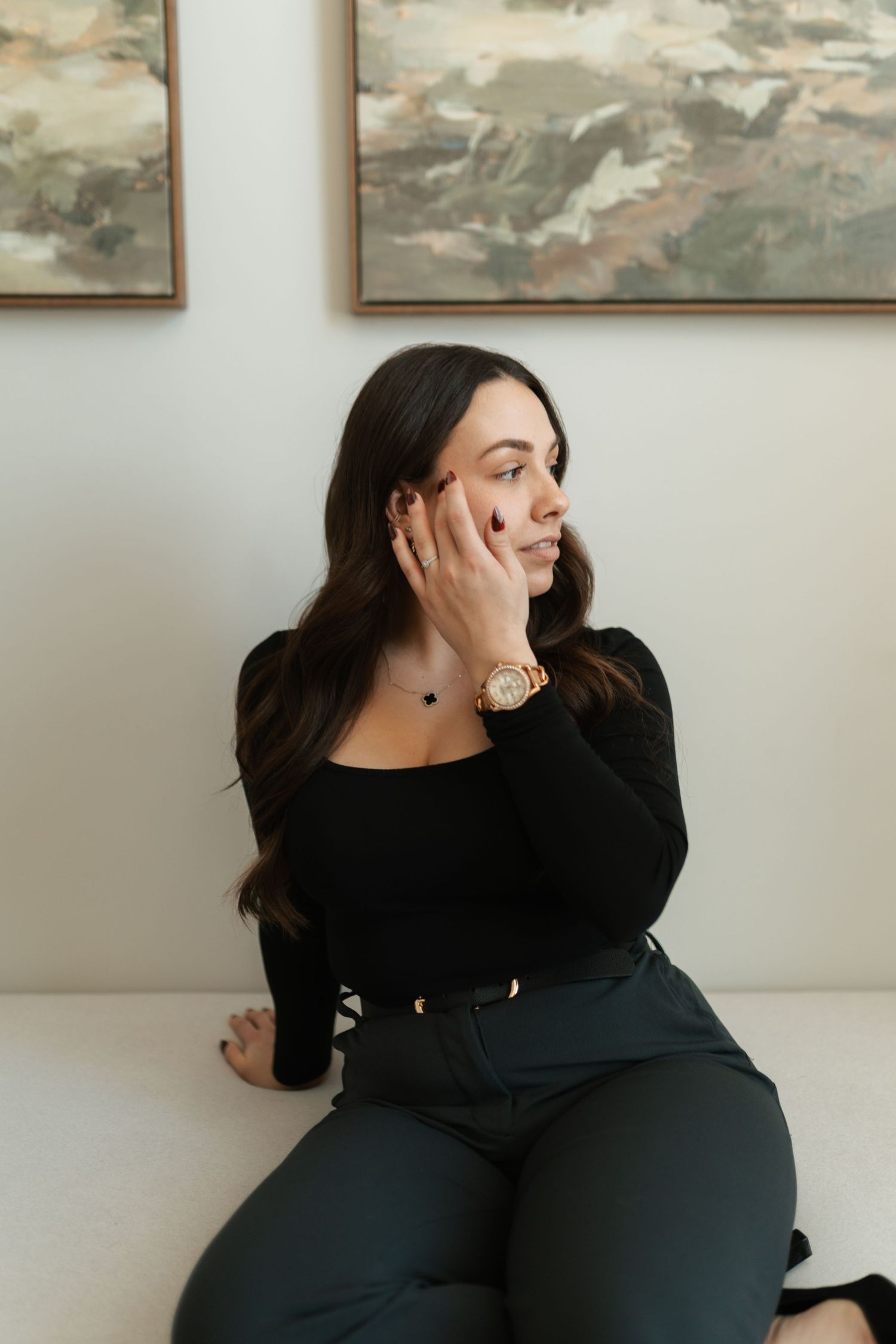 Brunette woman in black outfit with gold watch sitting on white couch beside abstract wall art.