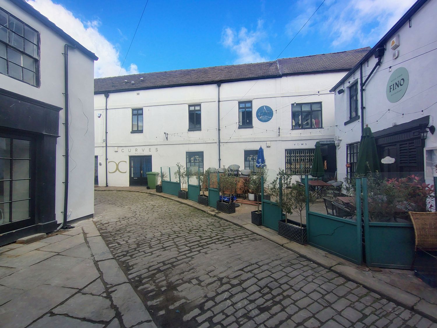 Cobblestone courtyard surrounded by white-painted buildings with green shopfronts and potted plants under a blue sky.