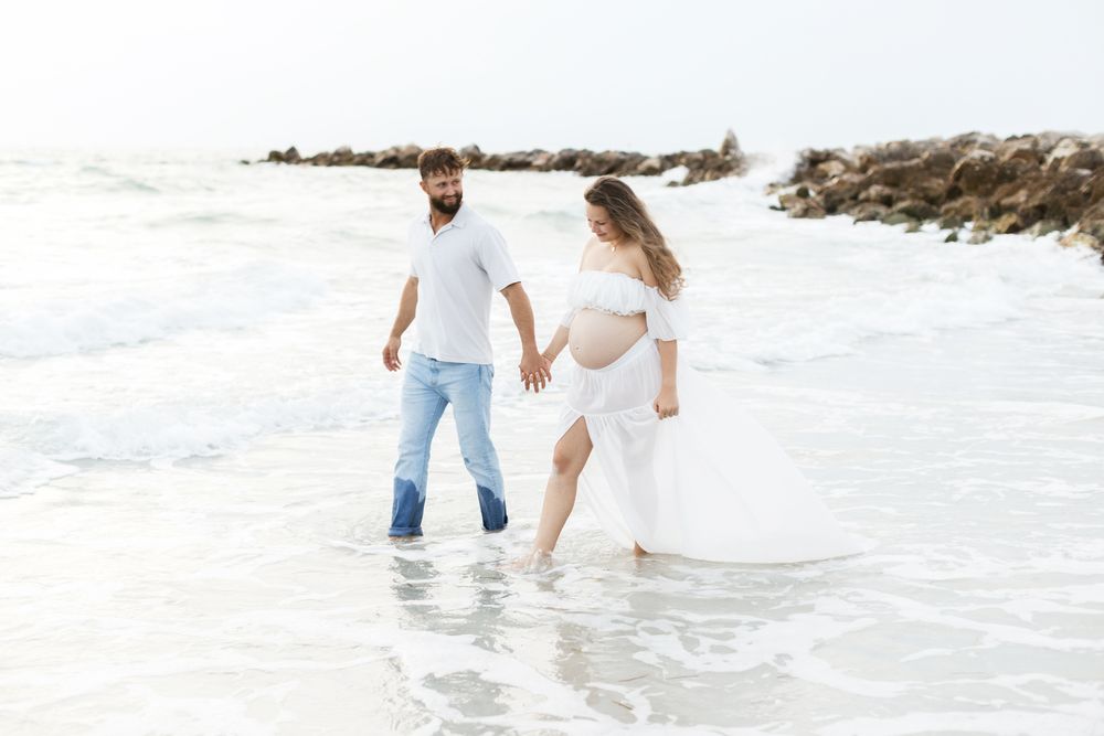Man in blue jeans holds expectant mother's hand wearing white outfit walking in shallow ocean water. Southern Maine maternity photographer