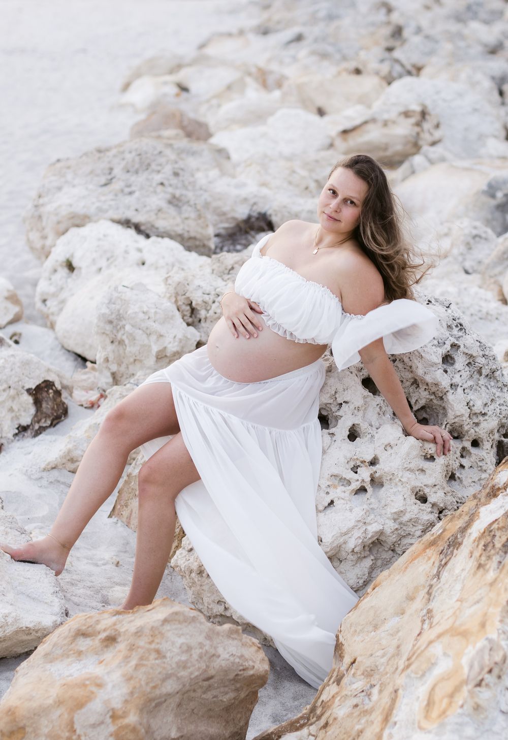Expectant mother wears white dress posing on light coastal rocks for maternity photos. North Shore Newburyport maternity photographer