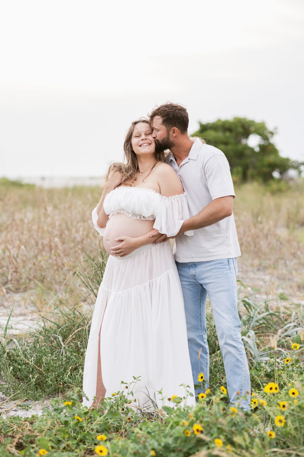Expectant mother wears sheer white outfit is snuggled from boyfriend standing in coastal grass for maternity photos. Maternity photographer