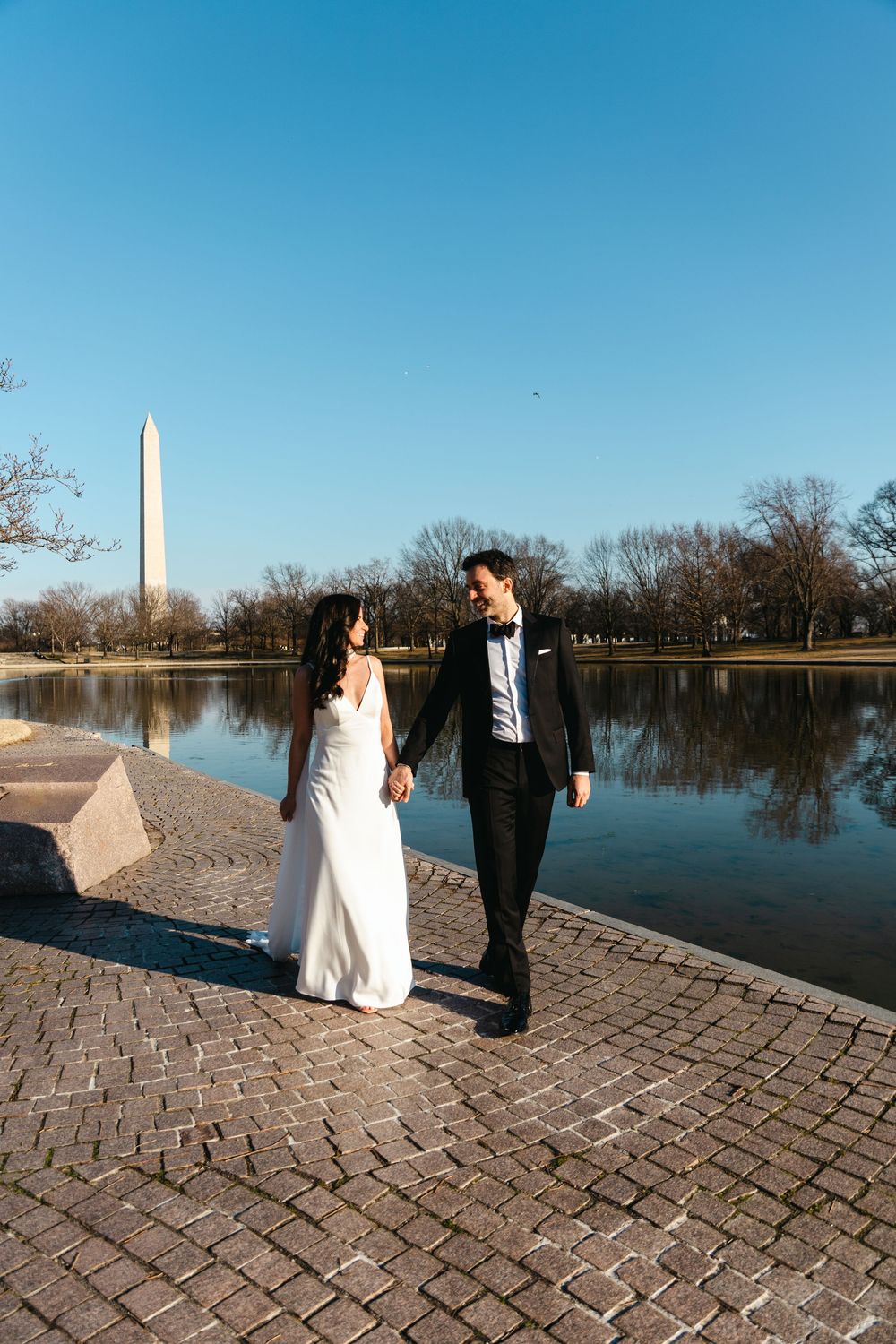 Bride and groom holding hands walking along a cobblestone path by the Washington Monument Reflecting Pool.