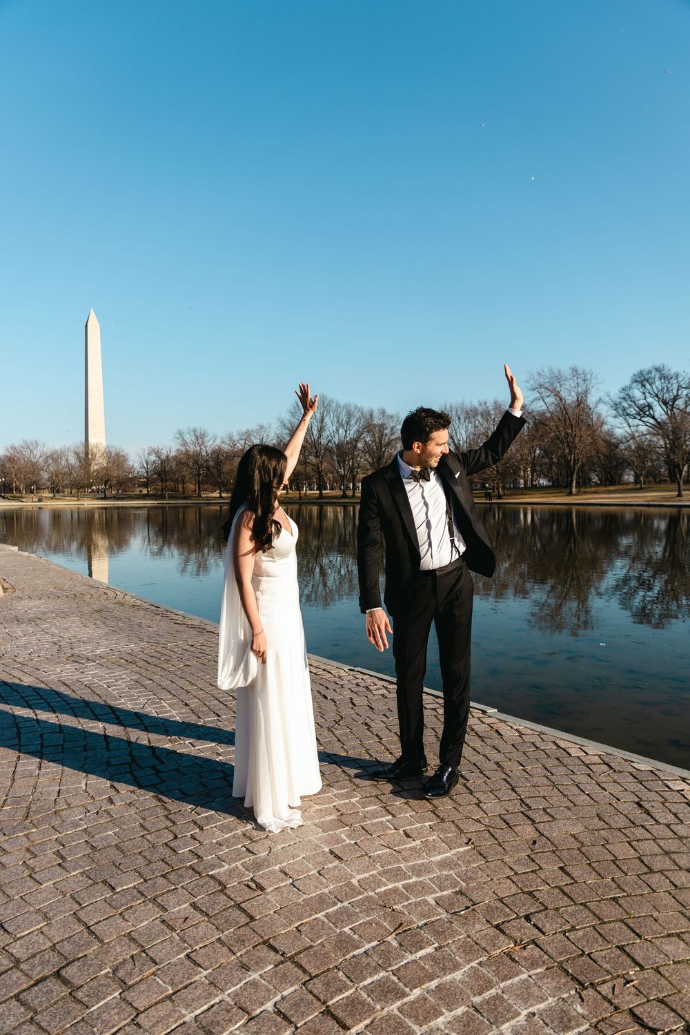 Bride and groom joyfully raising their arms by the Washington Monument Reflecting Pool on a sunny day.