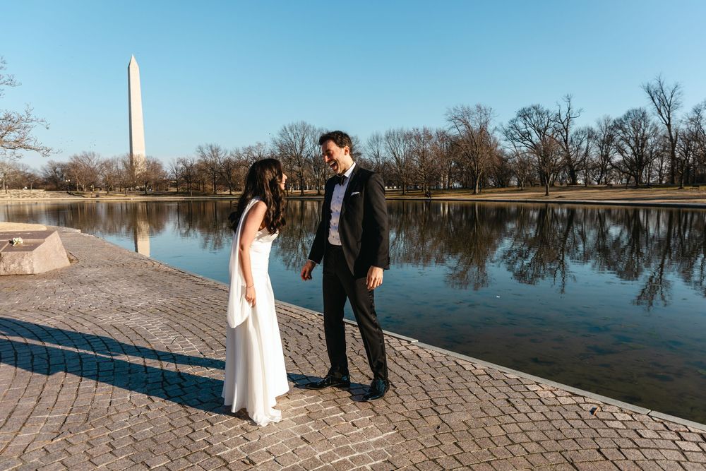 Couple in wedding attire holding hands by the Tidal Basin with Washington Monument reflected in water.