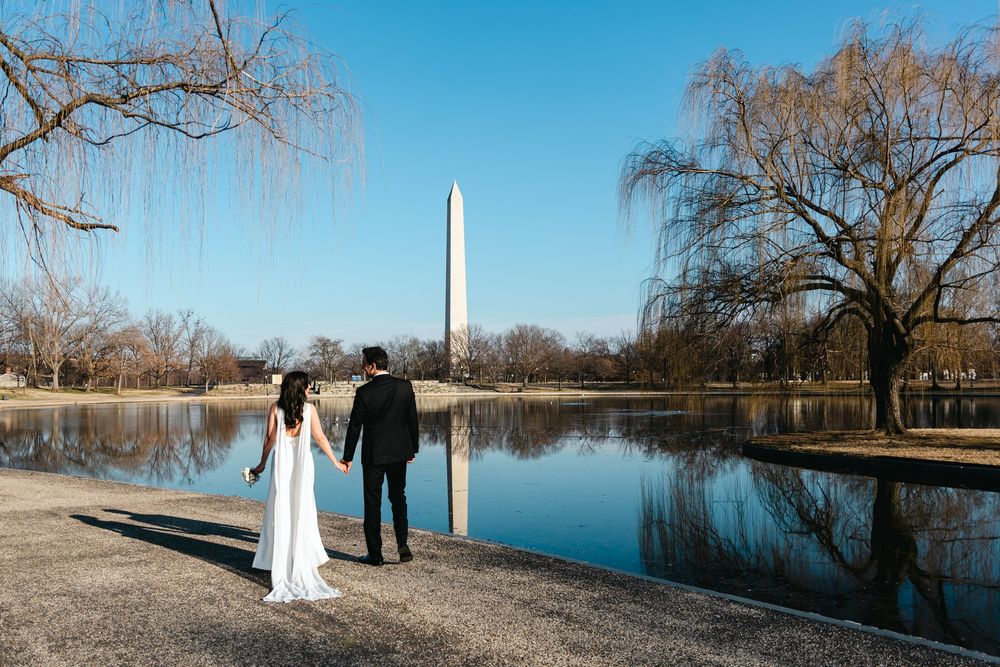 Bride and groom holding hands facing the Washington Monument reflected in the Tidal Basin on a clear sunny day.