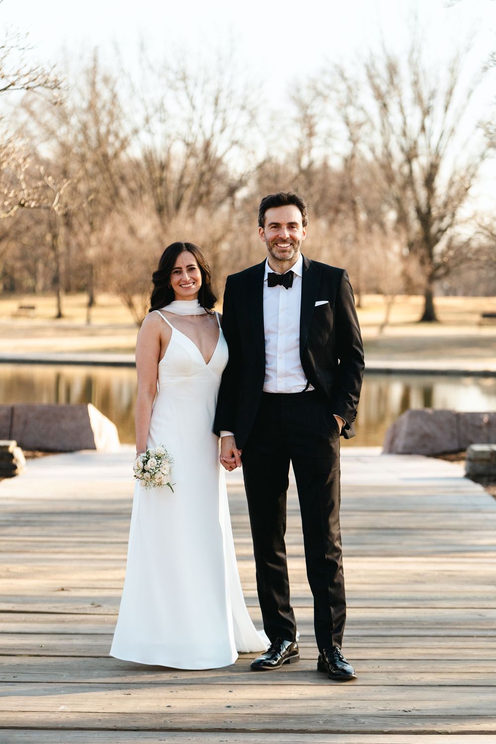 Bride and groom posing on a wooden dock near a lake, she in a white gown, he in a black tuxedo.