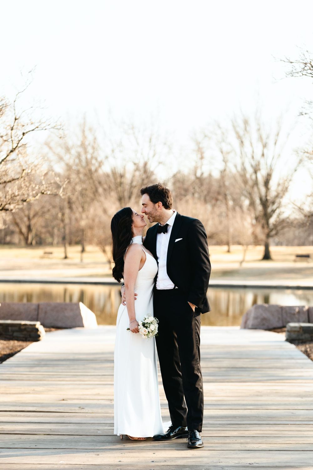 Bride and groom share a romantic moment on a wooden dock by a lake, she in a white gown, he in a black tuxedo.