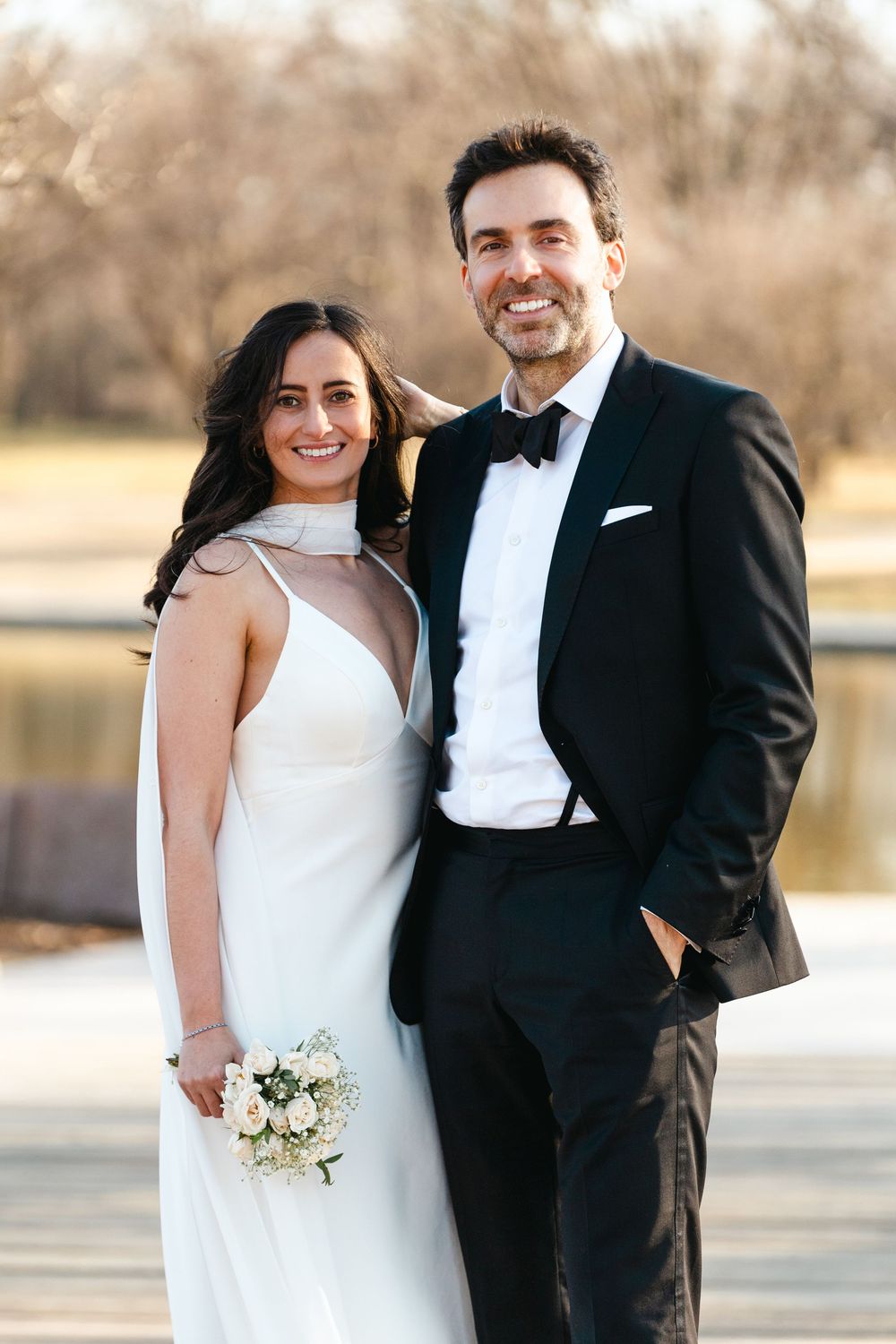 Smiling bride and groom posing together outdoors, bride holding a white bouquet in an elegant halter wedding gown.