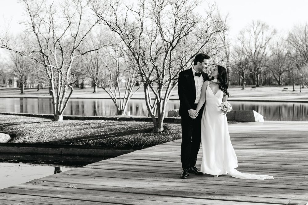 Black and white wedding photo of bride and groom kissing on a dock by a serene pond surrounded by bare trees.